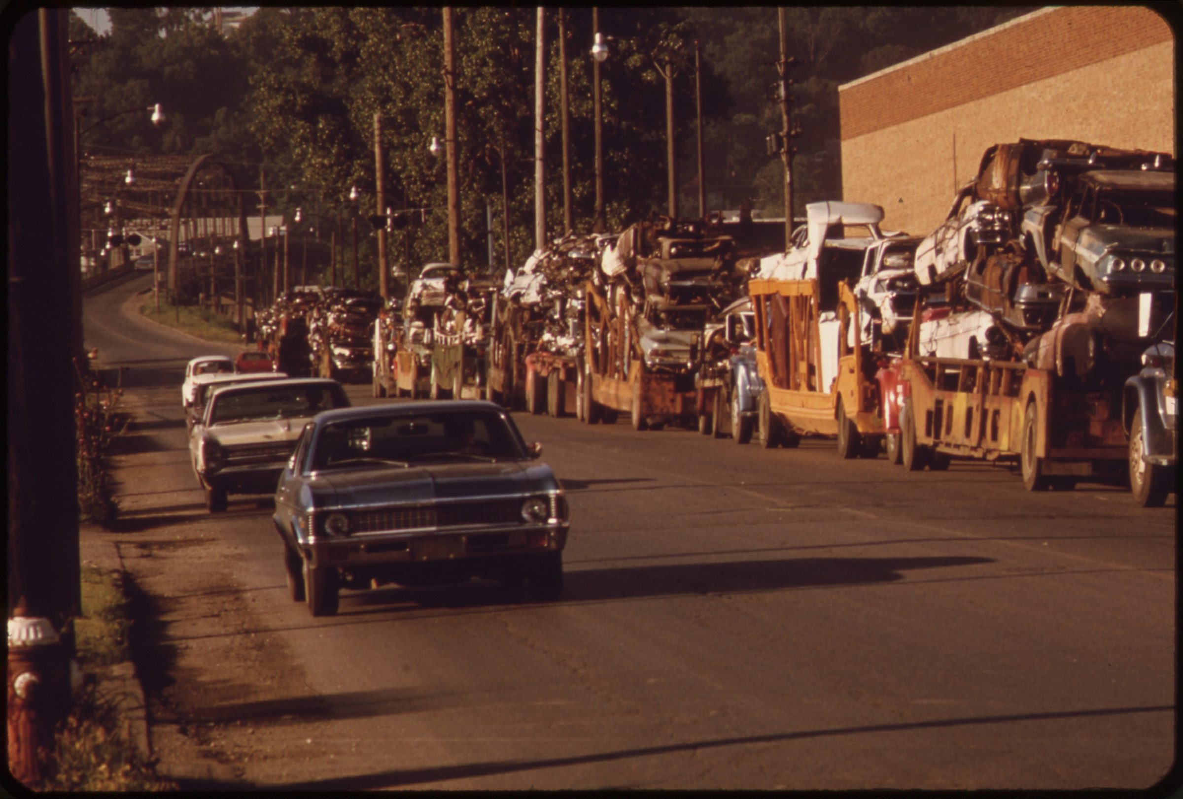 Twelfth Street at dawn on color film, car haulers stacked with junked cars lining the curb while one blue sedan drives through low golden light.
