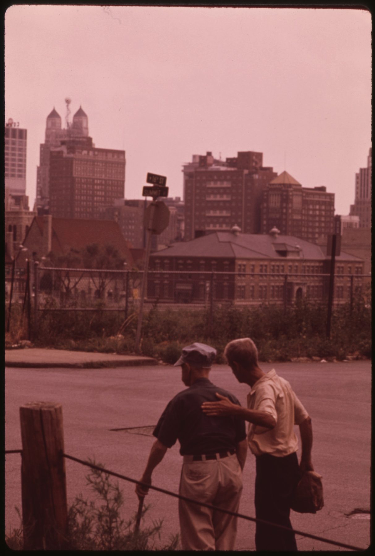 Two older men seen from behind at a low guardrail on an empty street, the downtown Kansas City skyline standing beyond the cleared ground in front of them.