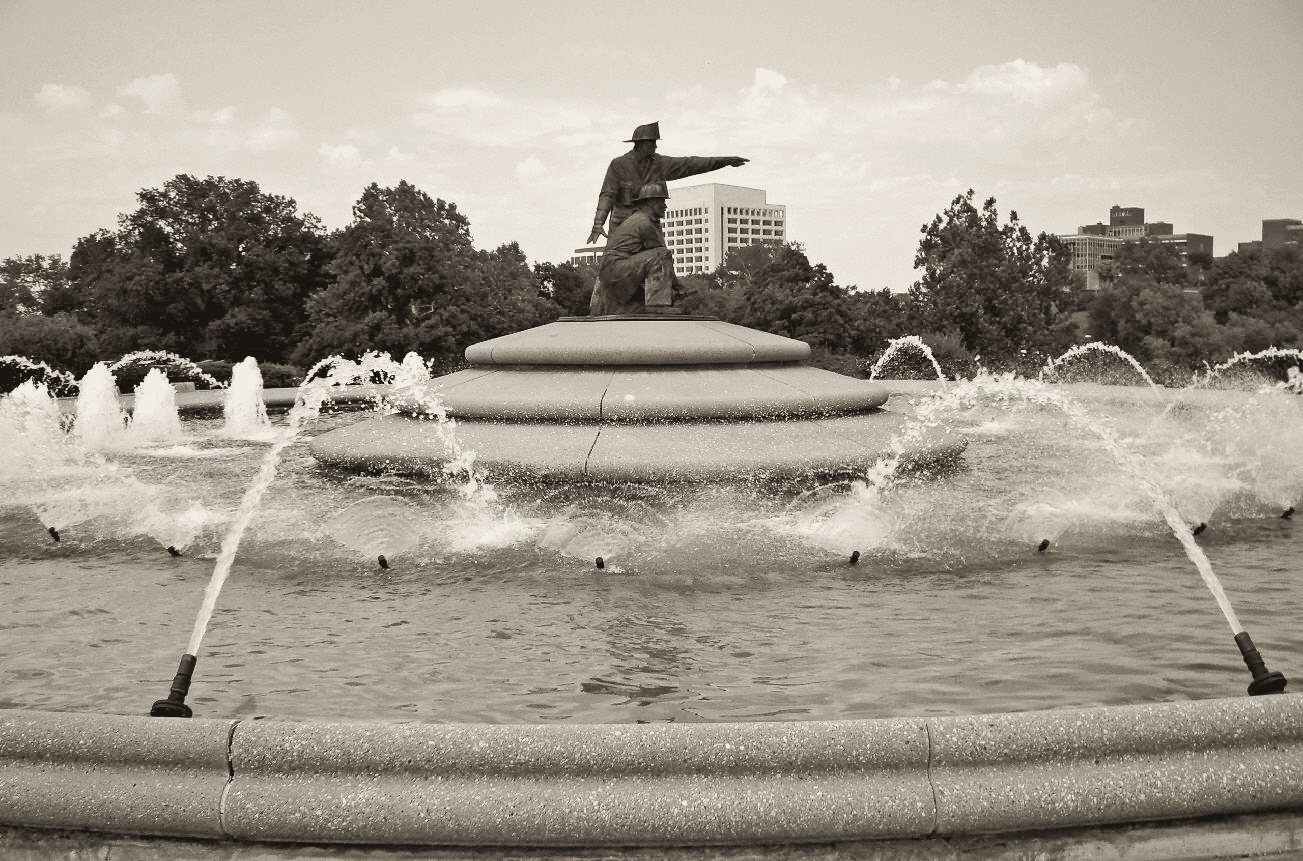 Warm-toned black and white print of the Firefighters Fountain, a raised bronze figure over rings of arcing water in Penn Valley Park.