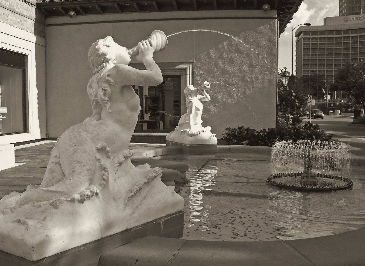 Warm-toned black and white print of a marble siren blowing a conch at the edge of a pool, a second figure standing in the spray behind her.