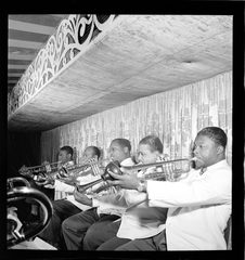A row of trumpet players seated shoulder to shoulder under a low decorated ceiling, bells up.