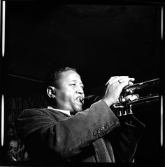 A trumpet player mid-phrase under one hard light against a black room.